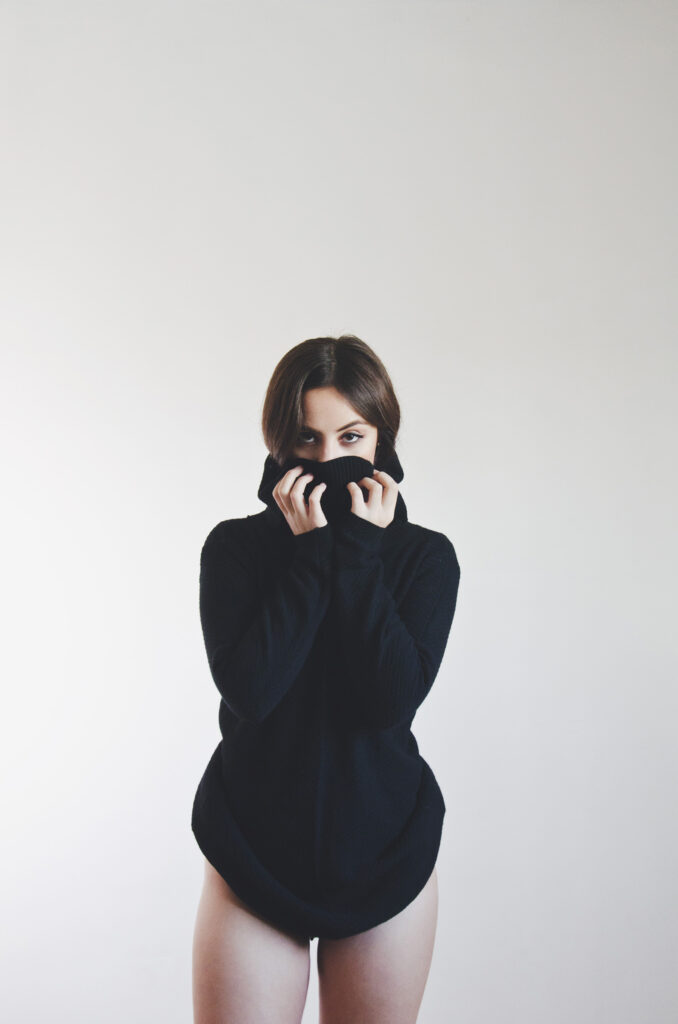 Laura Adriana photo by Ivan Beoulve. A brunette in a black turtleneck sweater stands against a white backdrop pulling the sweater up to her nose revealing only her eyes.